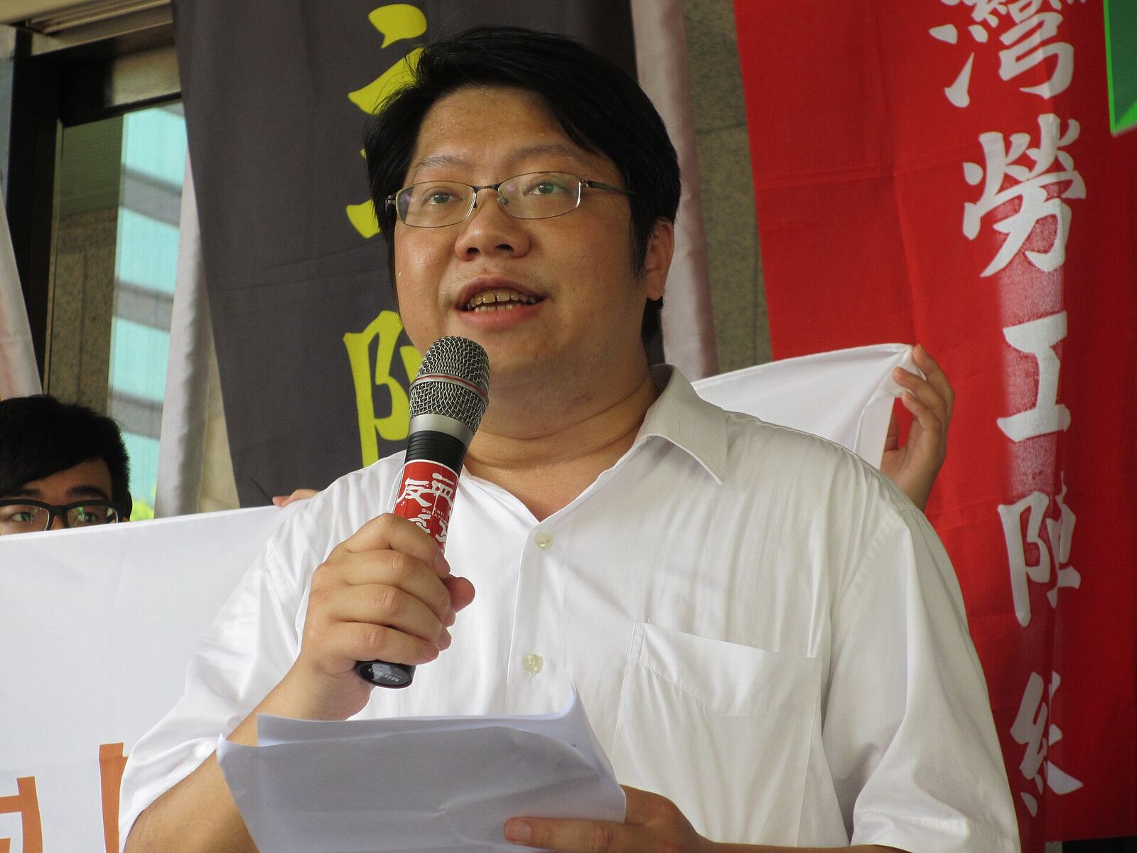 A man speaks into a microphone at a political rally, representing labour rights advocacy.
