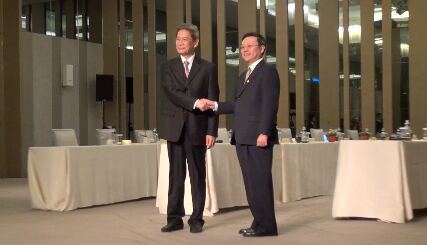 Two men shake hands at an indoor business event, demonstrating diplomacy.